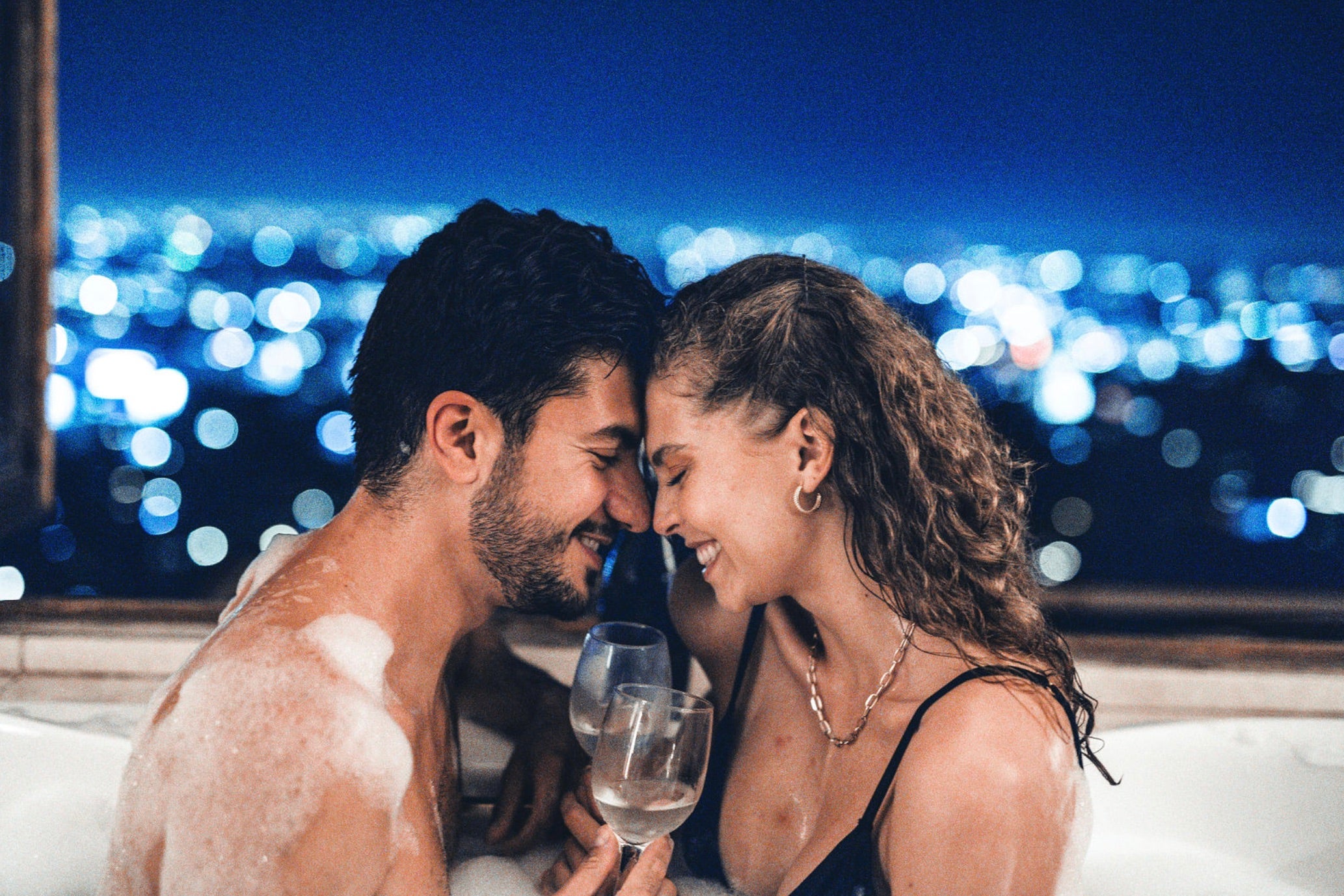 Couple press their foreheads against each other and drink champagne while taking a bath together