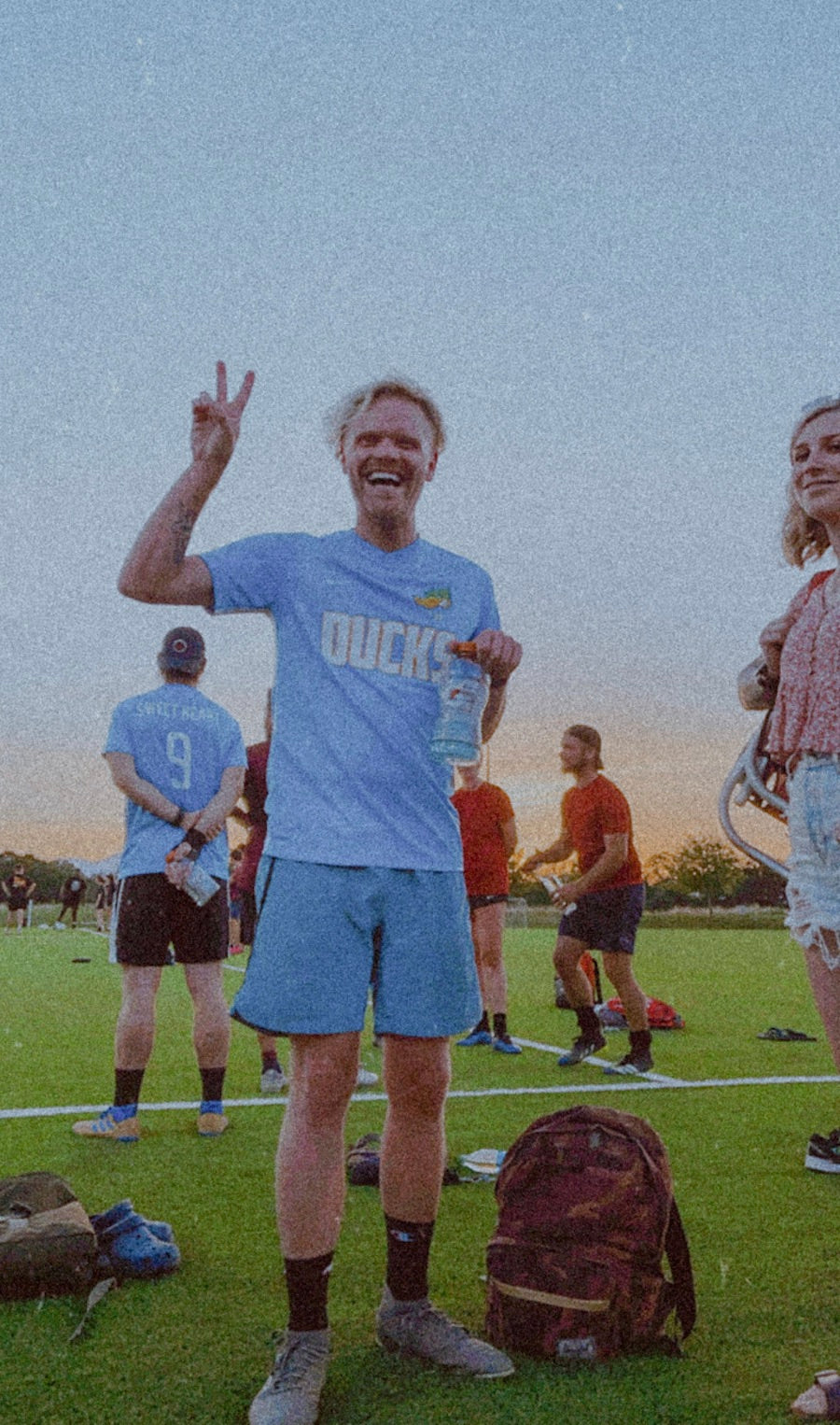Guy making a peace sign as he plays soccer with his friends at an outdoor field.