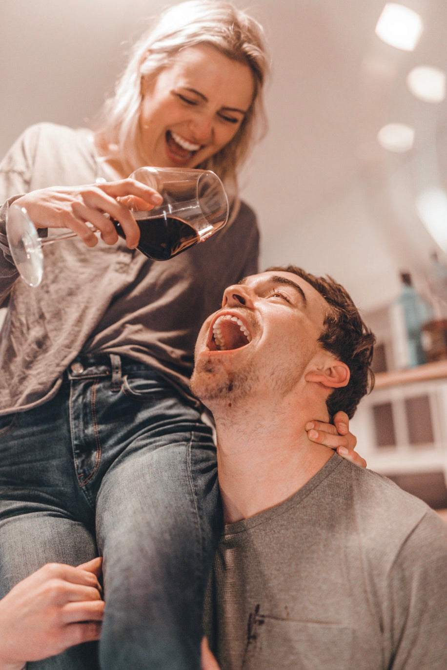 Wife sitting on husband's shoulder and playfully making him drink wine from above