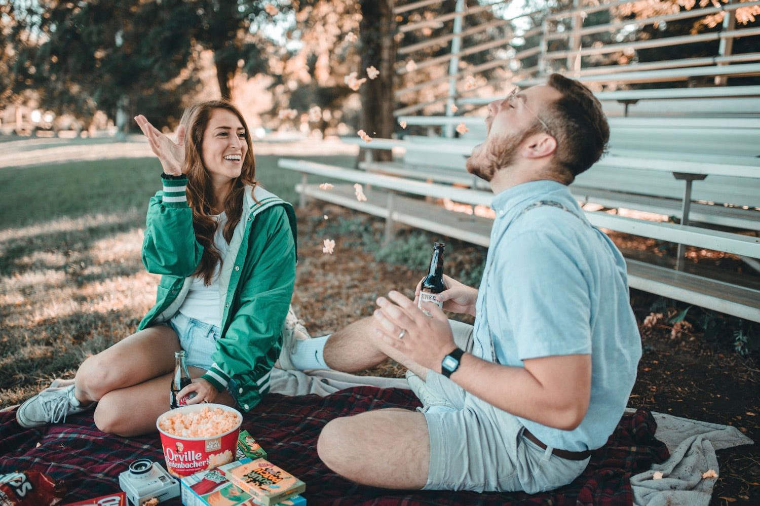 An image of a girl throwing snacks into her boyfriends mouth as they have a picnic together.