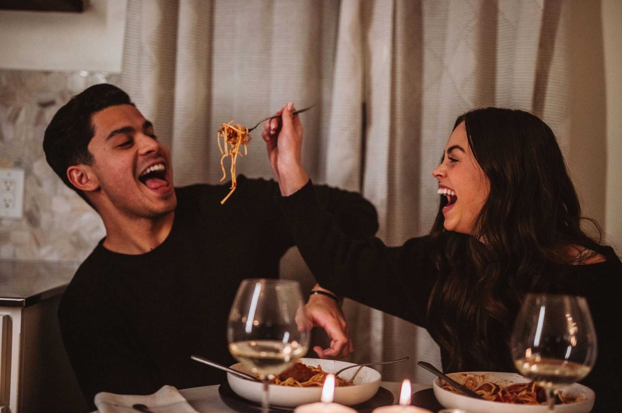 At Home Date Night Ideas: Couple sharing laughs as they feed each other spaghetti during their dinner date