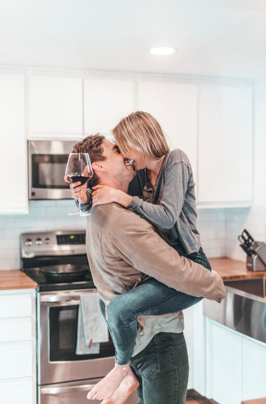 Husband carries wife who is carrying a glass of wine as they both smile in their kitchen