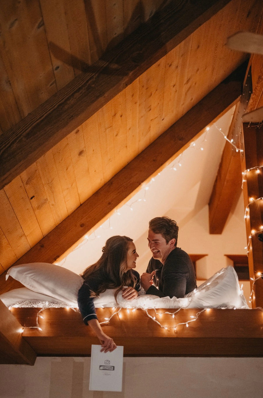 Couple smiling as they lay in bed with fairy lights, with the girl holding The Adventure Challenge Book