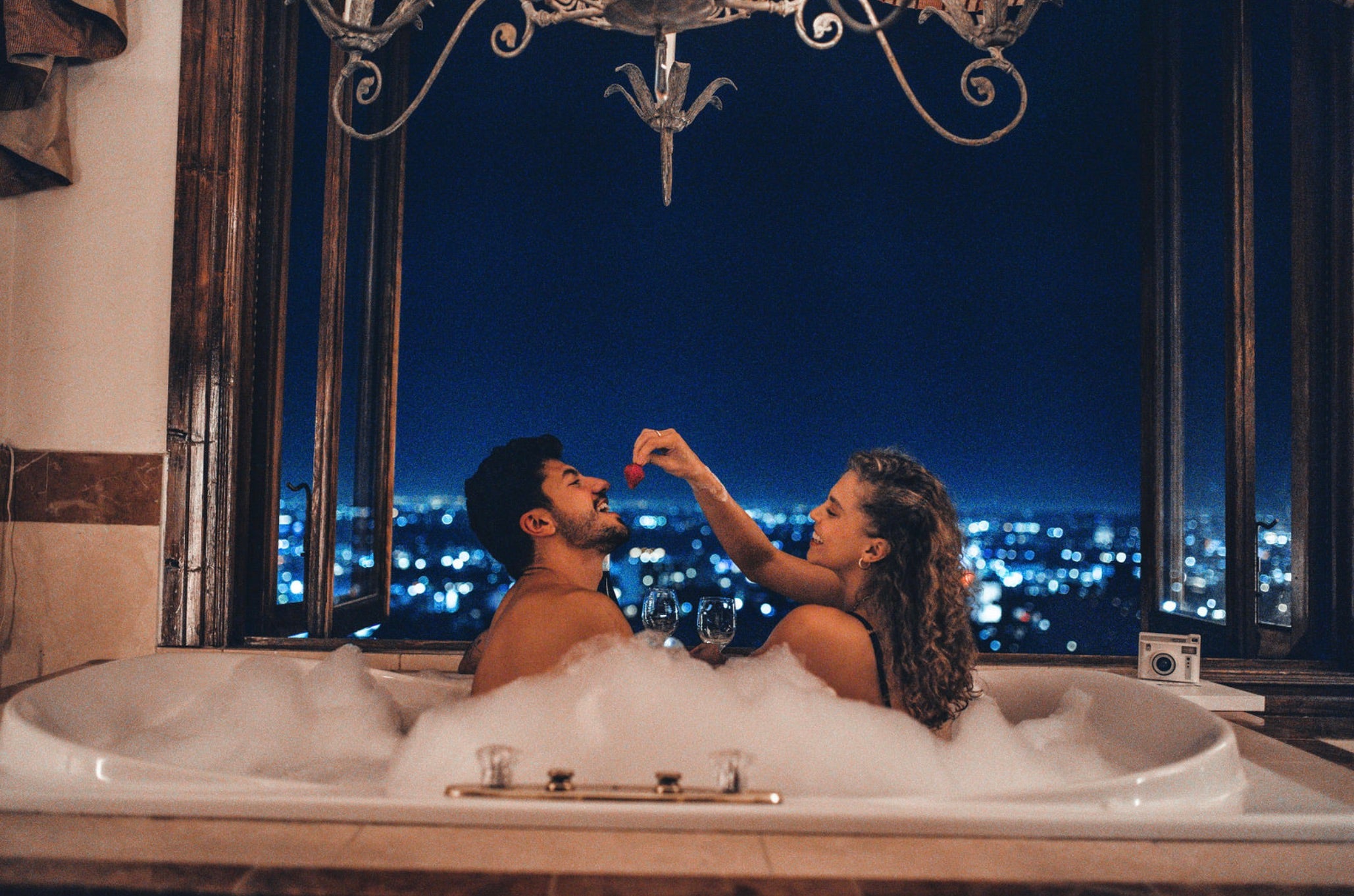 Girlfriend feeds her boyfriend a strawberry while in a bubble bath with a view of the city lights in front of them