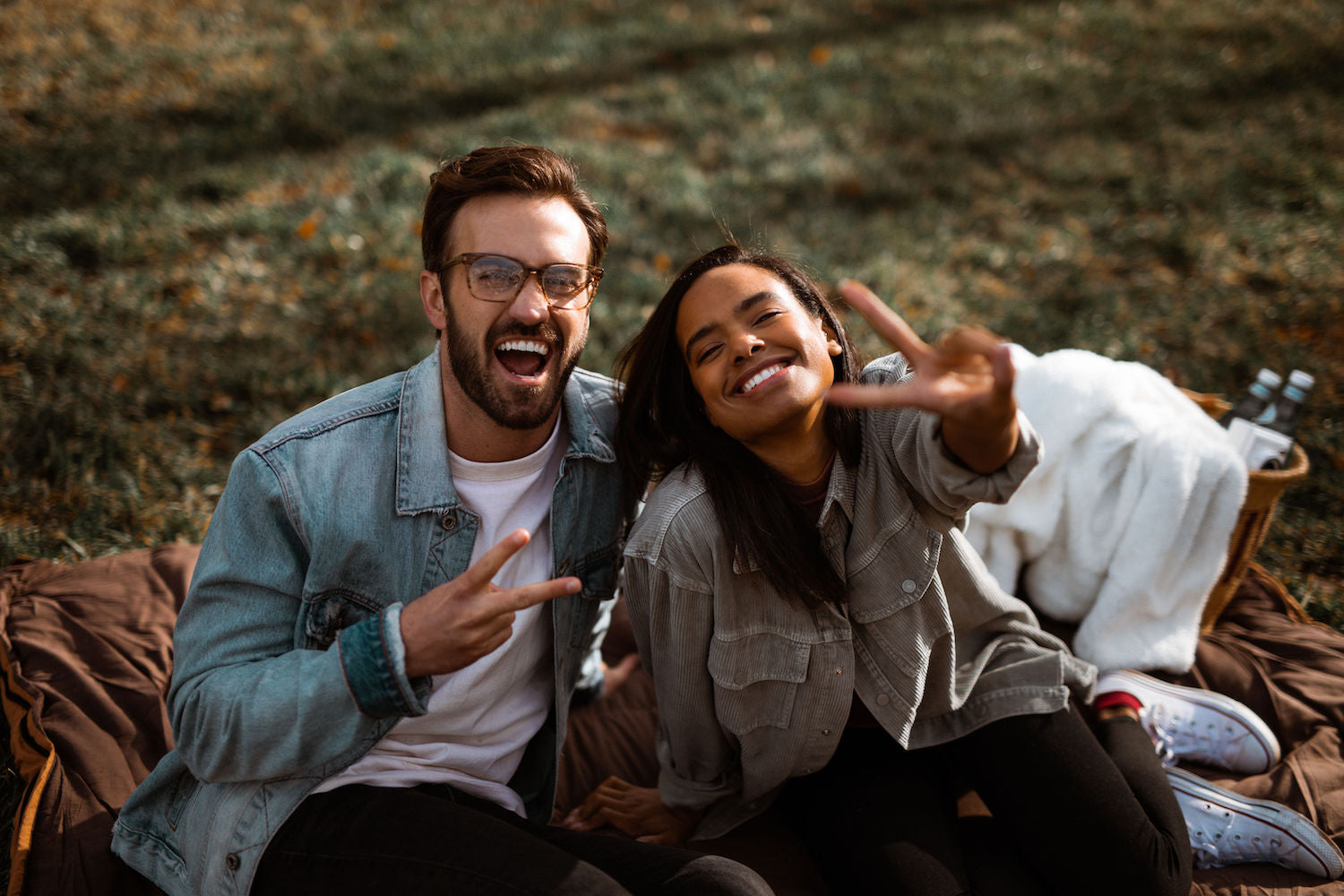 A couple sits on a picnic blanket smiling and making peace signs