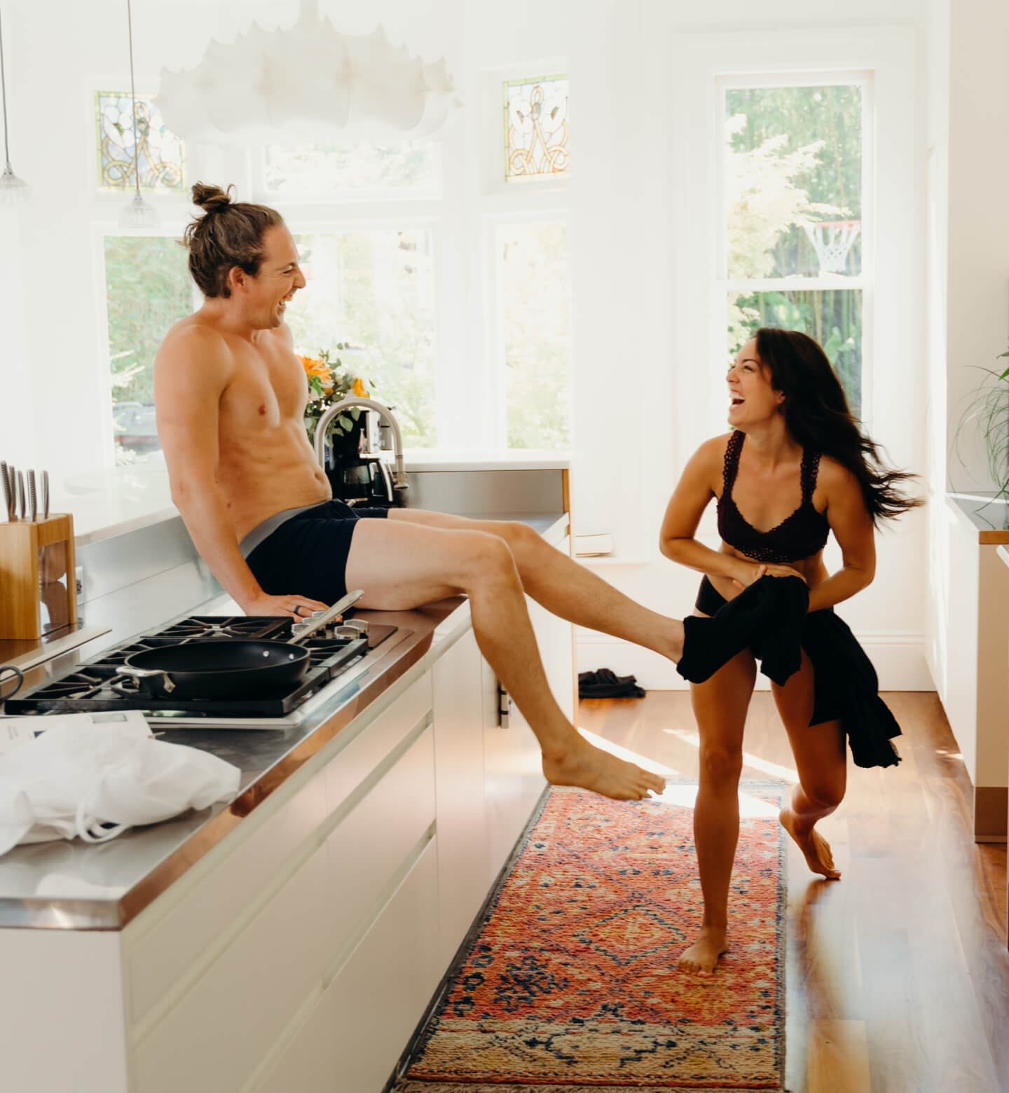 a man sits on the counter and laughs as his wife is being silly and taking off his pants